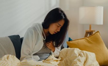 A woman with long black hair is sitting up in bed wincing in pain and one hand is on her chest.